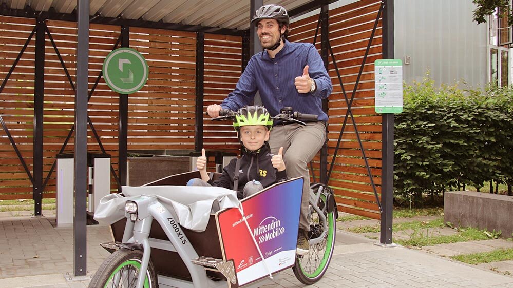 Ein Mann sitzt an einem Fahrrad, welches eine eingebaute Kiste vor dem Lenkrad beinhaltet. In dieser sitzt ein Junge. Beide tragen einen Helm und zeigen den Daumen nach oben.
