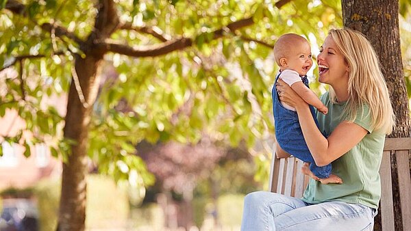 Eine Frau sitzt auf einer Bank im grünen und hebt ein Baby vor sich hoch. Beide wirken glücklich.